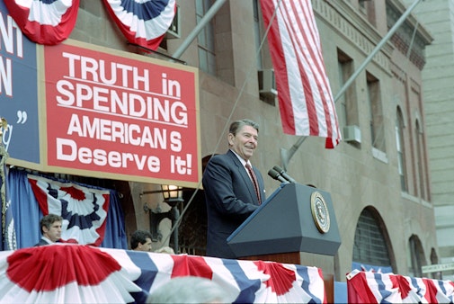 President Reagan speaking at a podium in front of brown building with red sign that reads "Truth in Spending American's Deserve It"