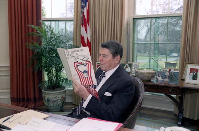 President Reagan sitting in the oval office reading a pamphlet